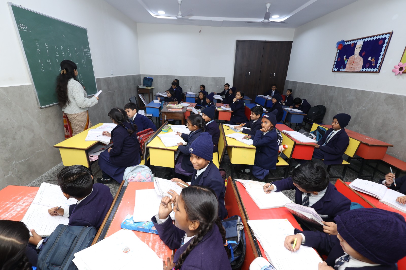 Teacher writing the alphabet on the blackboard while children watch from yellow desks