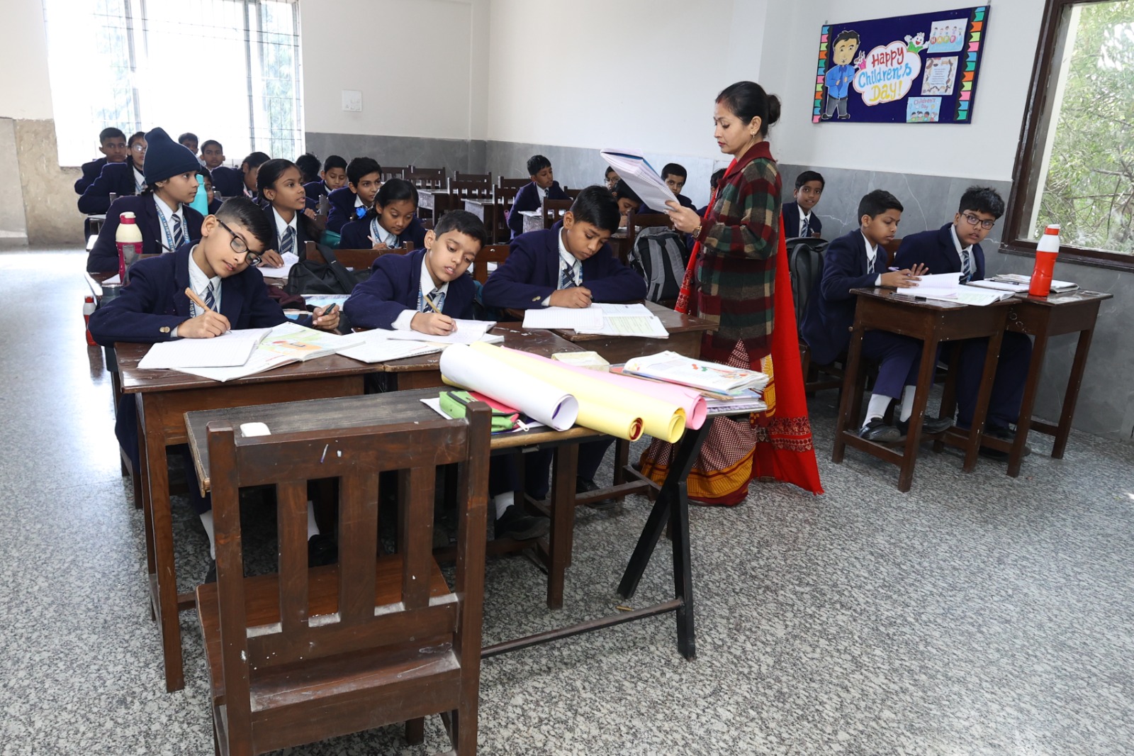 Senior students at wooden desks while a teacher reads aloud from a notebook