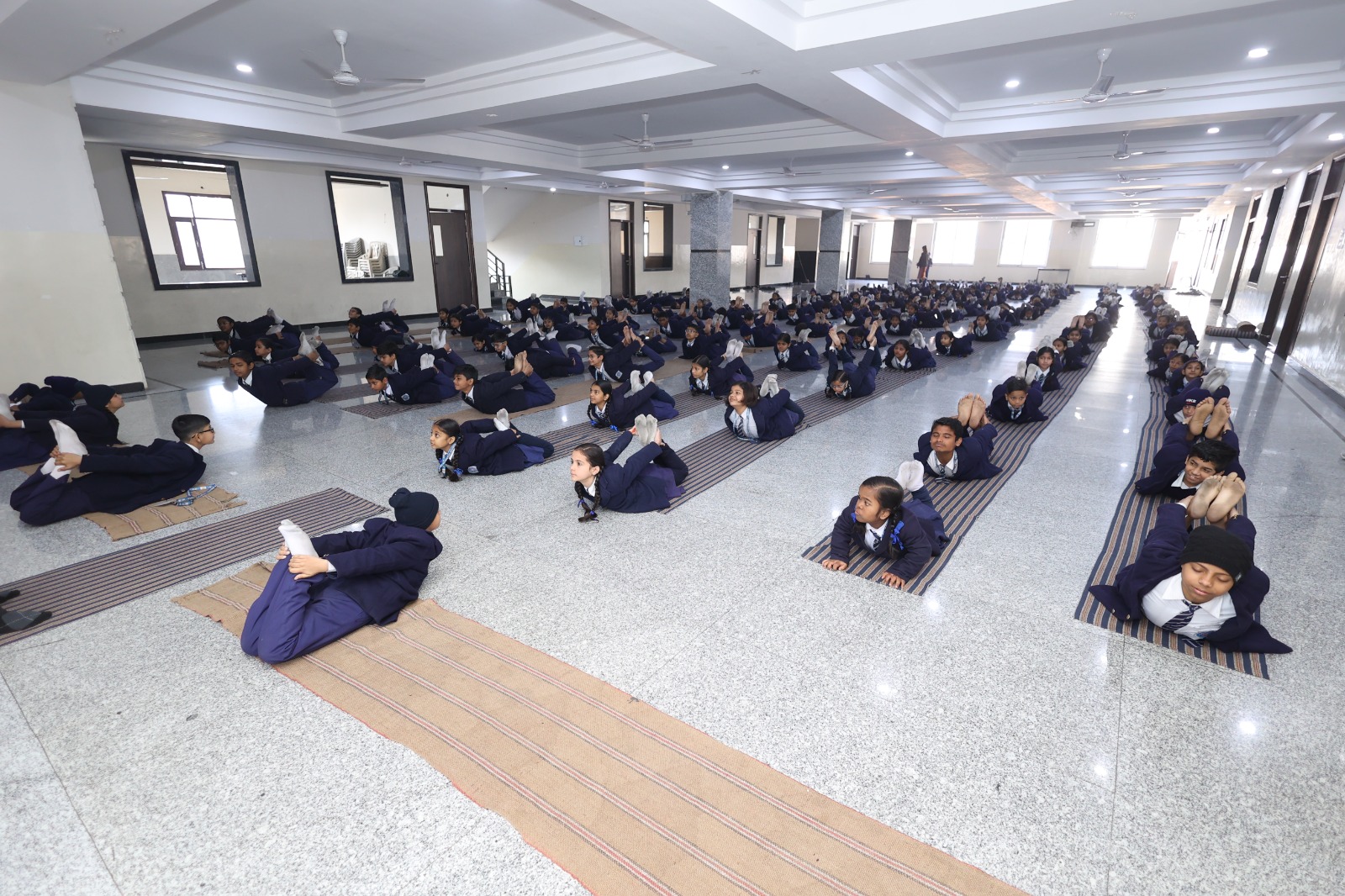 Indoor yoga hall with students lying on mats and a teacher walking down the centre